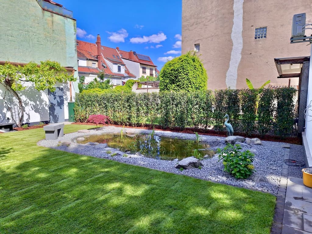 Angelegter Gartenteich mit Kiesufer und Findlingen, eingefasst von Rasenfläche und Kirschlorbeerhecke