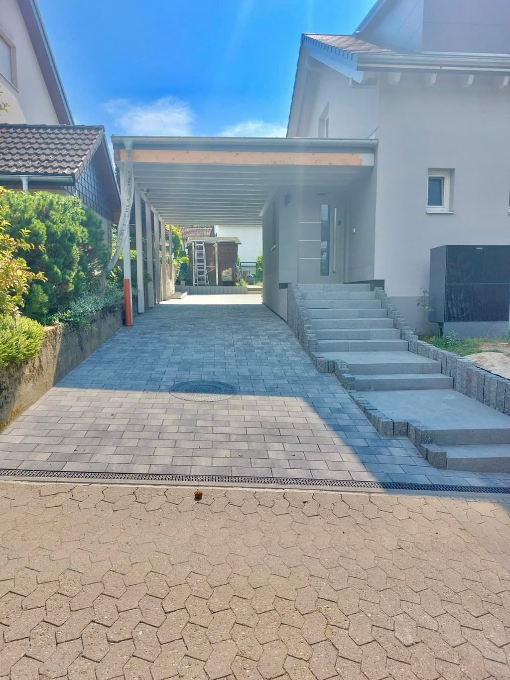 Gepflasterte Einfahrt unter einem Carport, daneben eine Blockstufentreppe zur Haustür